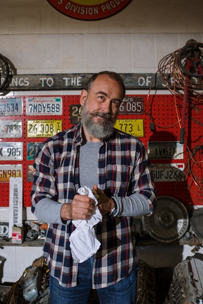 Bearded mechanic standing in a garage surrounded by license plates and car parts.