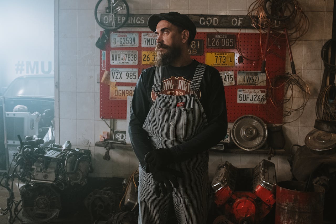 Bearded mechanic standing in a vintage garage with license plates and car parts.