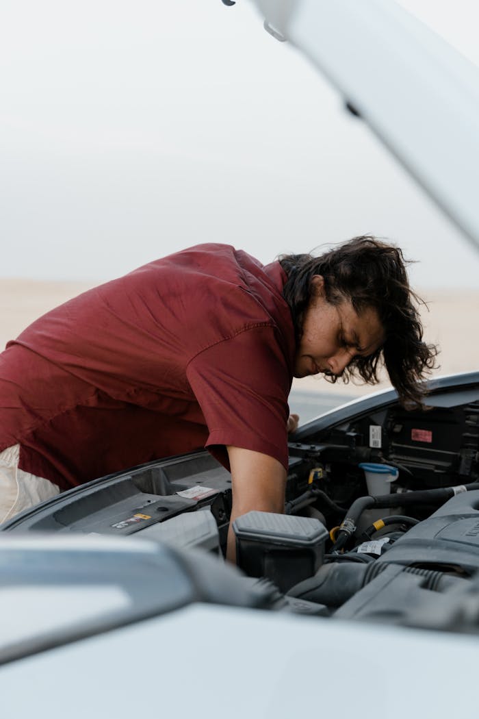 A man with long hair repairing a car engine in a desert setting, showcasing mechanics and adventure.